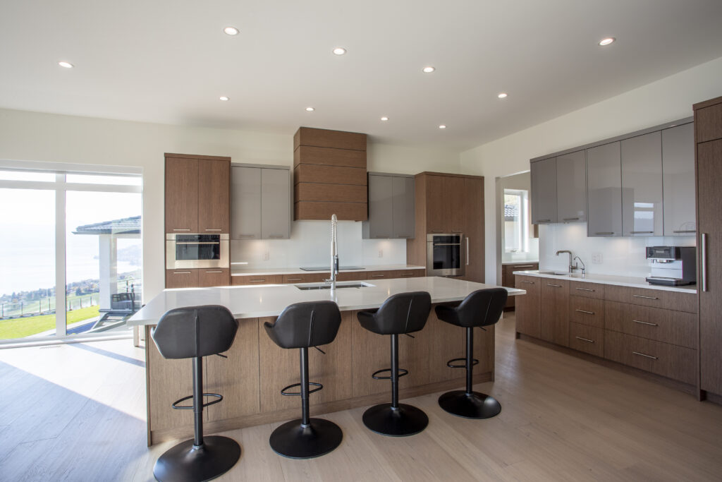 Modern kitchen with wood cabinetry, a large island with bar stools, and ample natural light from large windows overlooking the outdoors.