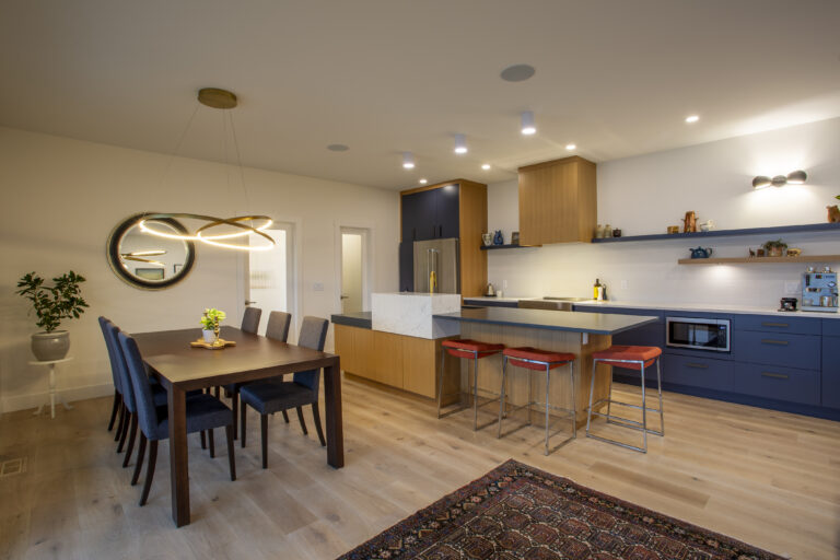 Modern open-concept dining area and kitchen with a sleek wooden dining table, contemporary lighting, and a spacious kitchen with a mix of blue and wood cabinetry.