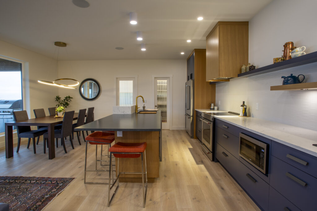 Full view of a modern kitchen with a central island, blue and wood cabinetry, and stylish lighting, seamlessly connecting to the open living and dining spaces.