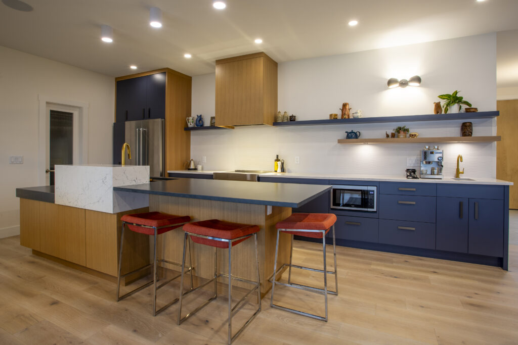 Modern kitchen featuring a central island with a marble countertop, red bar stools, blue cabinetry, and open wooden shelves displaying decor items, with sleek lighting and stainless steel appliances.