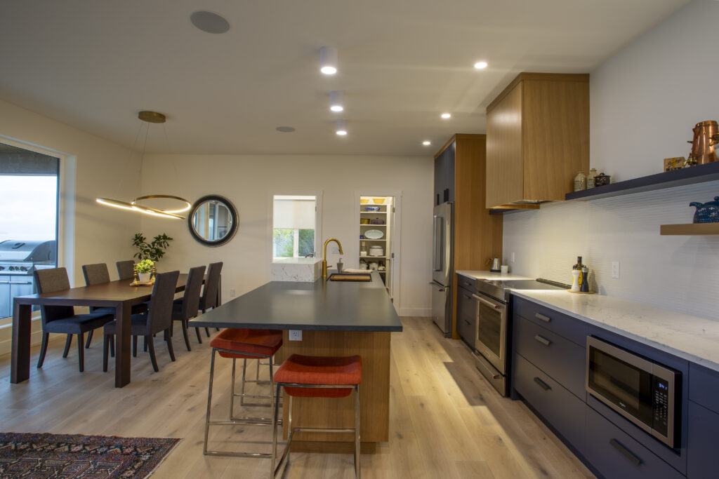 Full view of a modern kitchen with a central island, blue and wood cabinetry, and stylish lighting, seamlessly connecting to the open living and dining spaces.