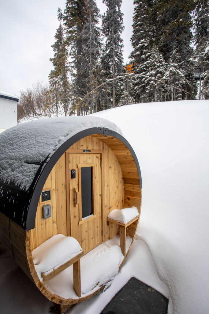 Outdoor barrel sauna surrounded by snow with a forest backdrop.