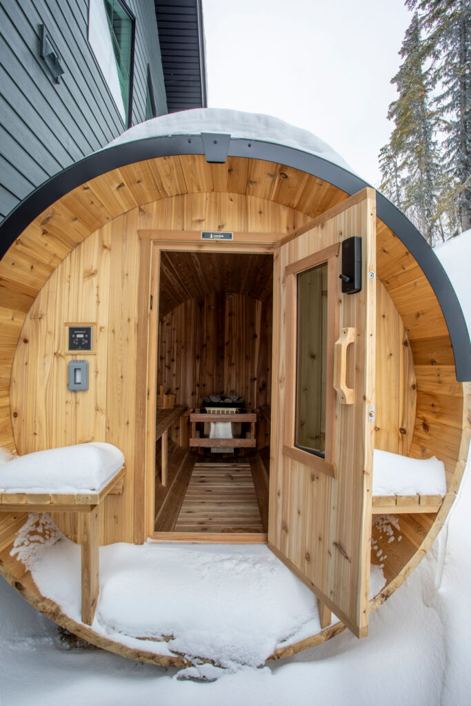 Outdoor barrel sauna surrounded by snow with a forest backdrop.