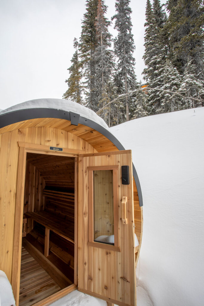 Outdoor barrel sauna surrounded by snow with a forest backdrop.