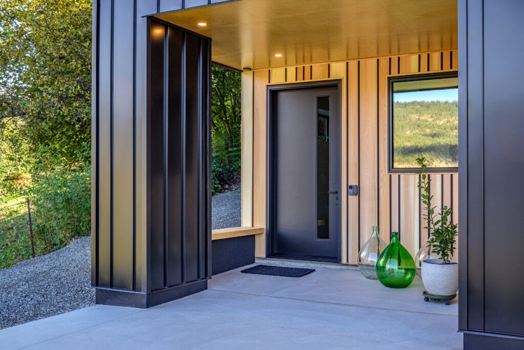 Sleek front entrance of a modern Lake Country home with wood and metal siding, a black door, and minimalist decor.