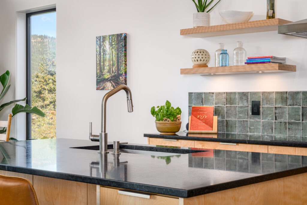 Kitchen sink with a black soapstone countertop, modern chrome faucet, open wood shelving, and a green tiled backsplash.