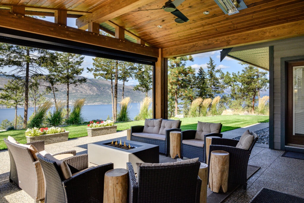 A covered outdoor lounge with a fire table and cushioned seating, overlooking Okanagan Lake.