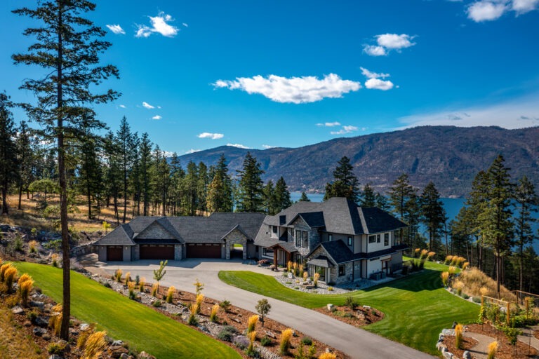 A Lake Country custom home with large windows, a winding driveway and a backdrop of Okanagan Lake and mountains.